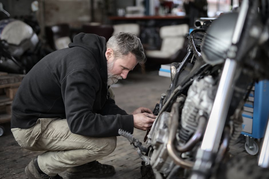 Side view of focused male mechanic in casual wear sitting near motorbike and examining details details in workshop